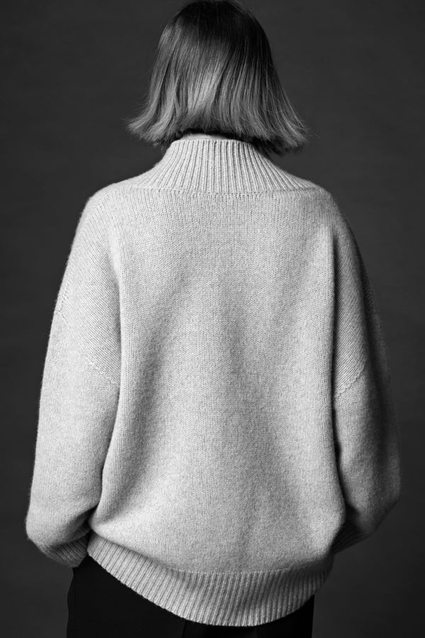 The back of a woman standing in a cosy v-neck cashmere sweater in light grey with her hands in her pockets.