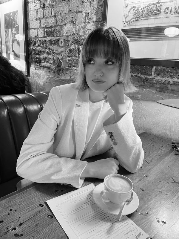 A woman sitting in a café wearing a horn-buttoned, cream-hued, single-breasted jacket and a T-shirt.