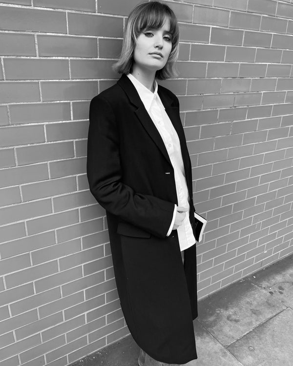 A young woman leaning against a wall, wearing a black, single-breasted coat, holding a note book in her left hand.