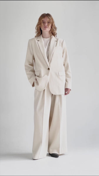 A video of a woman turning around in a cream-hued, wide-legged pair of trousers with pleats putting her hands in side slit pockets, styled as a suit with a matching single-breasted jacket and a white T-shirt. The mid-weight fabrics in the slightly loose-cut suit flow when she moves.