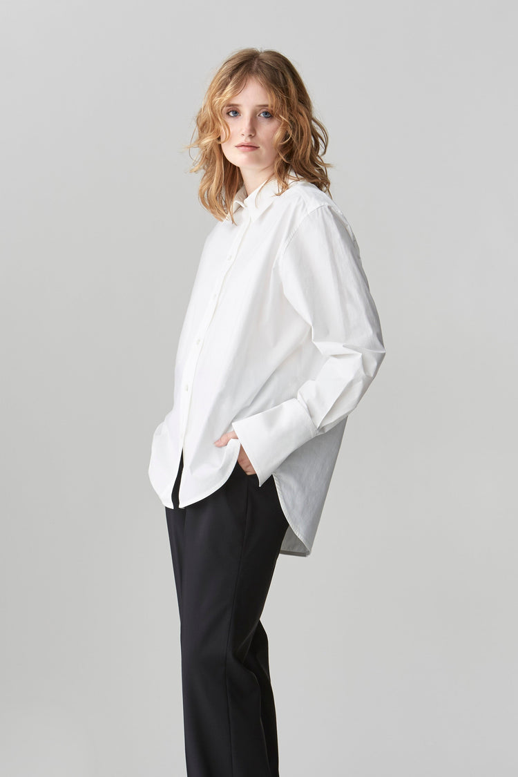 A woman wearing a crisp cotton poplin shirt with a high-low curved shirttail hem and deep cuffs with her hand in her black trouser pocket styled with black shoes.