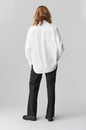 The back of a woman wearing a crisp cotton poplin shirt with a low curved shirttail hem and deep cuffs with her hands in her black trouser pockets styled with black shoes.