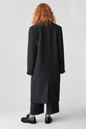 The back of a woman wearing a black coat with black horn buttoned sleeves and a back vent, styled with black trousers and black leather shoes.