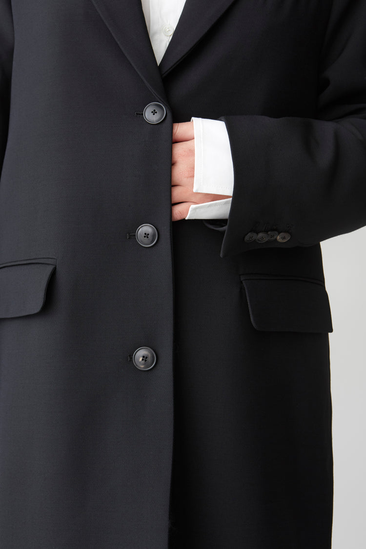 Details of a black horn buttoned, black coat front and sleeve with a crisp poplin shirt cuff sticking out serving a hint of rebellious attitude.