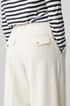 Details of the back of a cream-hued, wide-legged pair of trousers with pleats, horn-buttoned back-pocket flaps and side slit pockets, styled with a striped long-sleeve T-shirt.